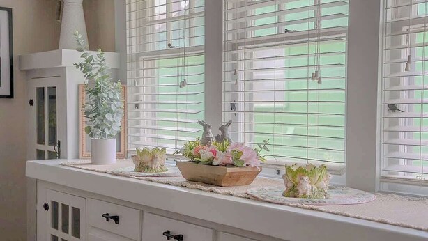 A white dresser with drawers and a mirror, a vase with flowers, and a window with blinds.