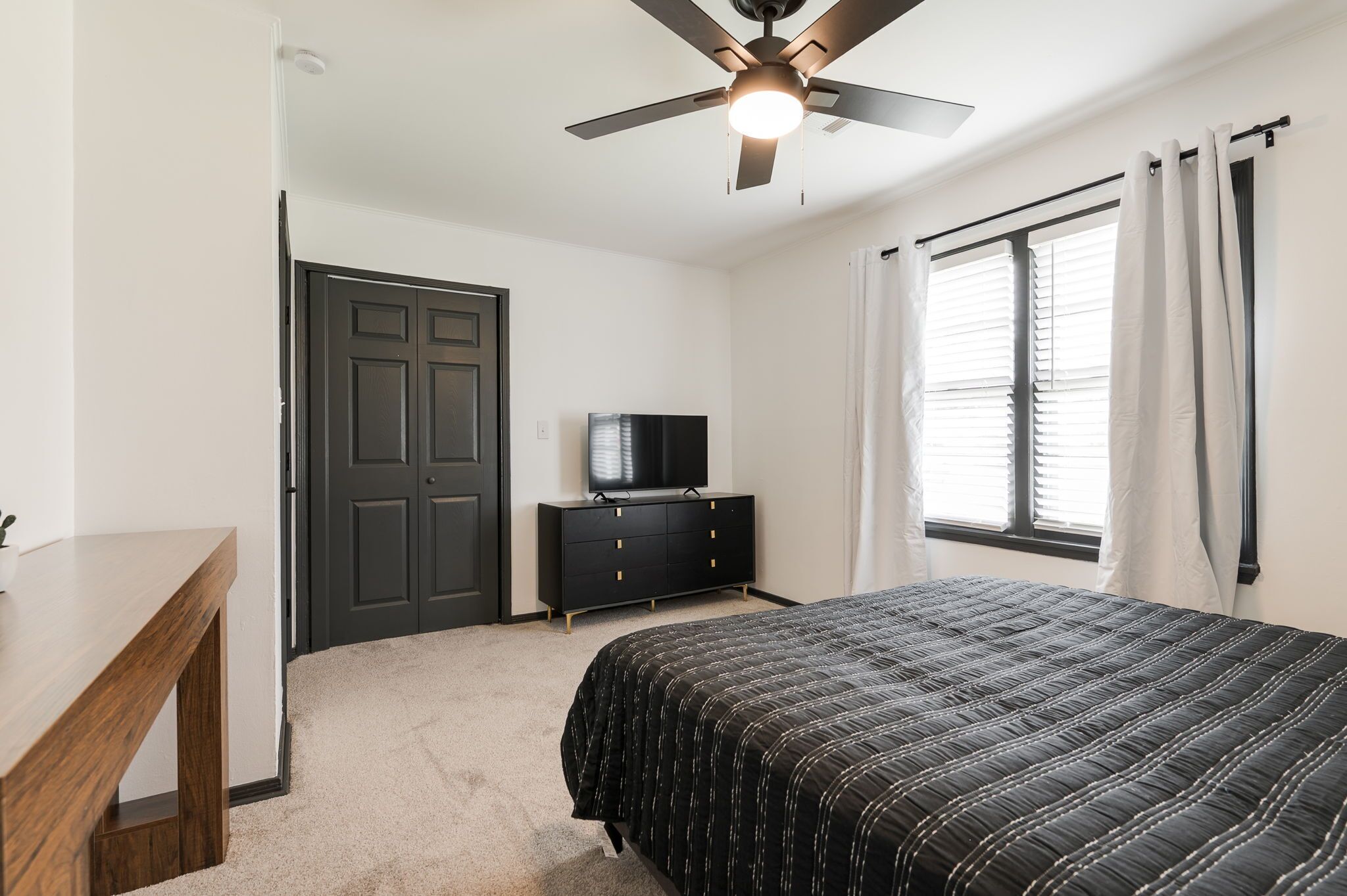 A bedroom with a bed, a dresser, a television, a ceiling fan, and a window with curtains.