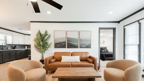 A modern living room with a brown leather sofa, two beige armchairs, and a wooden coffee table. There is a large potted plant and three framed artworks on the wall.