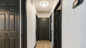 A narrow hallway with wooden flooring, a black door on the left, a black cabinet with drawers in the center, and a white wall with a clock on the right.