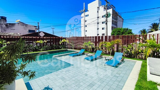 A swimming pool with lounge chairs, surrounded by greenery and a fence, with a building in the background. - Maré Alta Beach Hall-Praia de Tamandaré (Tamandaré)