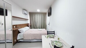 A hotel room with a bed, a bedside table with a remote control, a TV mounted on the wall, and a dining table set with plates and cutlery. - Maré Alta Beach Hall-Praia de Tamandaré (Tamandaré)