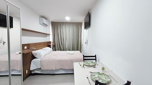 A hotel room with a bed, a bedside table with a remote control, a TV mounted on the wall, and a dining table set with plates and cutlery. - Maré Alta Beach Hall-Praia de Tamandaré (Tamandaré)