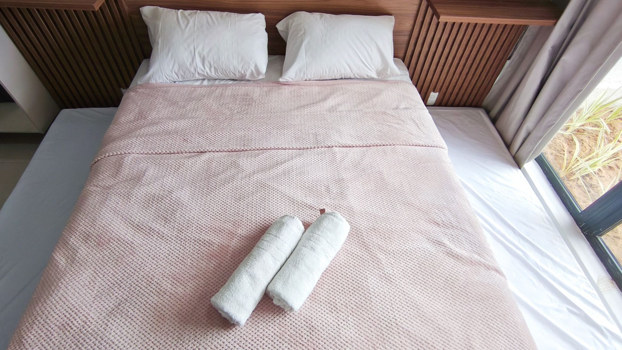 Uma cama com um edredom rosa texturizado, duas toalhas enroladas e almofadas brancas.