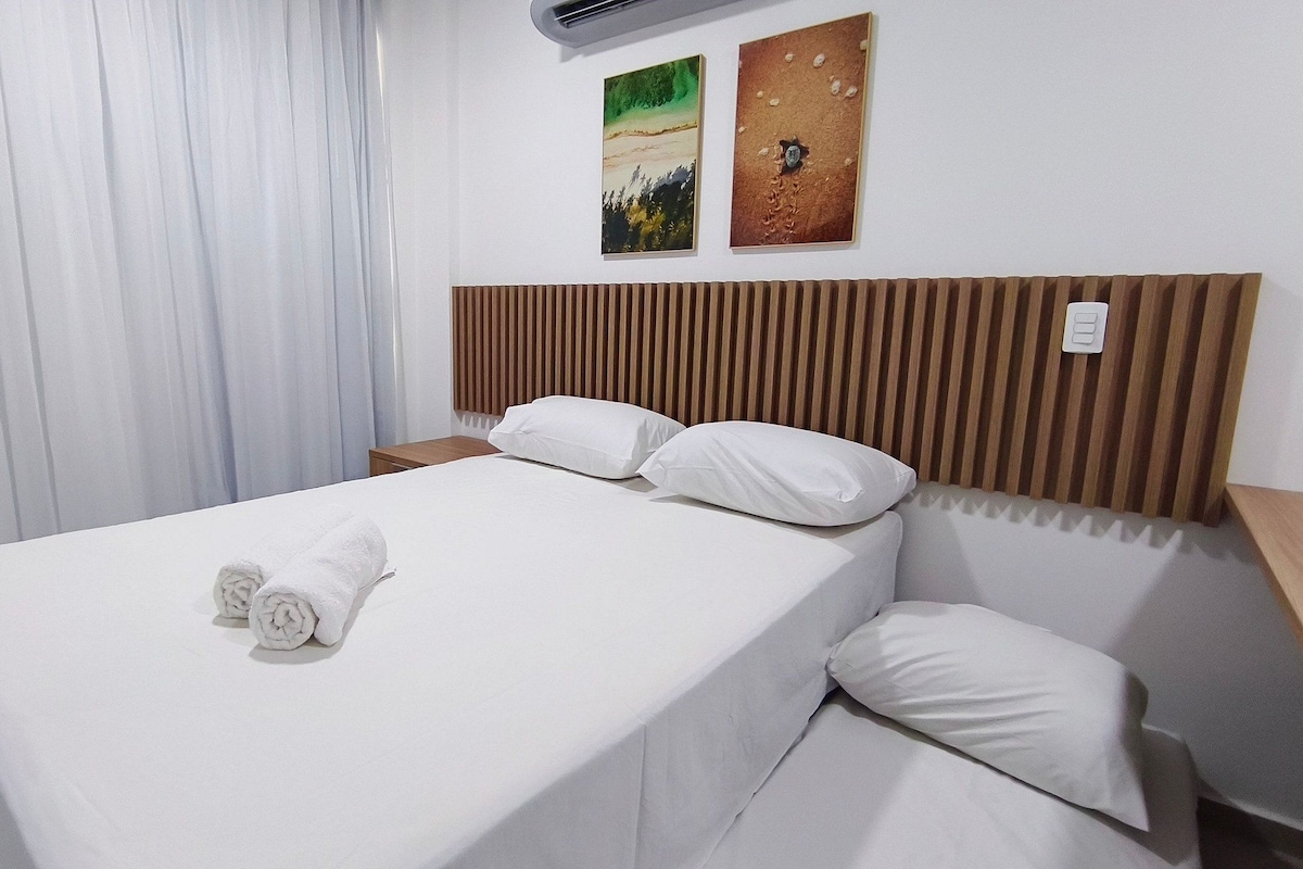 A neatly made bed with white linens and pillows, a wooden headboard, a nightstand, and two wall art pieces.