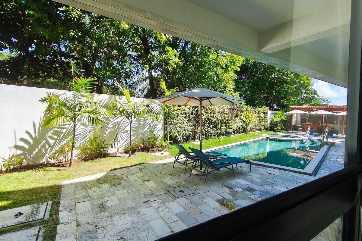 A swimming pool with lounge chairs, a pergola with an umbrella, and a view of a lush garden and building in the background.
