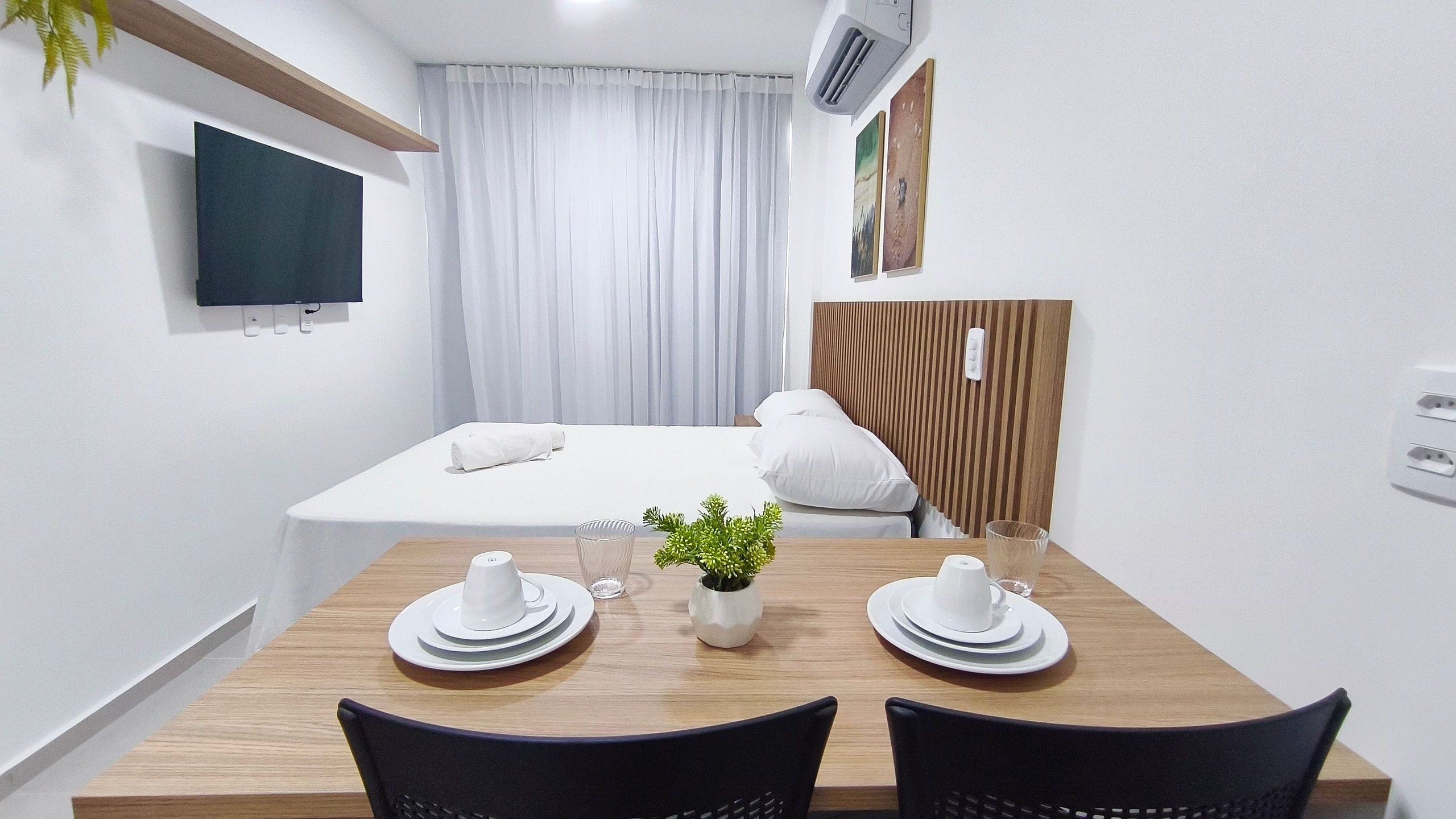 A hotel room with a bed, a TV mounted on the wall, a wooden table with two chairs, and a small plant on the table.