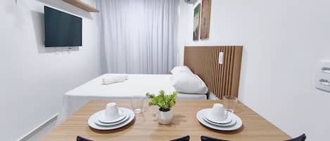Quarto de hotel com cama, televisão presa na parede, mesa de madeira com duas cadeiras e uma pequena planta sobre a mesa.