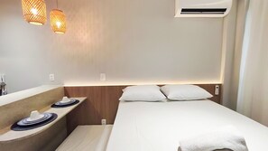 A hotel room with a bed, a bedside table with a lamp, an air conditioning unit, and a mirror.