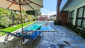 A swimming pool with lounge chairs and umbrellas, surrounded by greenery and a paved patio.