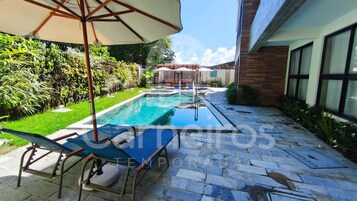 Piscina con sillones de descanso y sombrillas, rodeada de vegetación y un patio empedrado.