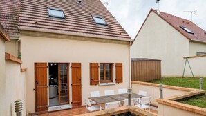 Ein zweistöckiges Haus mit einer Terrasse, einem Holztisch und Stühlen sowie einem kleinen Garten.