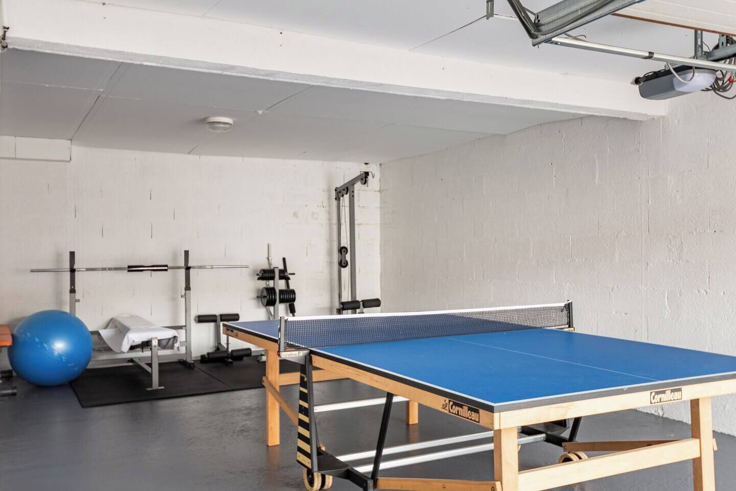 A ping pong table in a room with a white wall, a blue exercise ball, and weightlifting equipment.