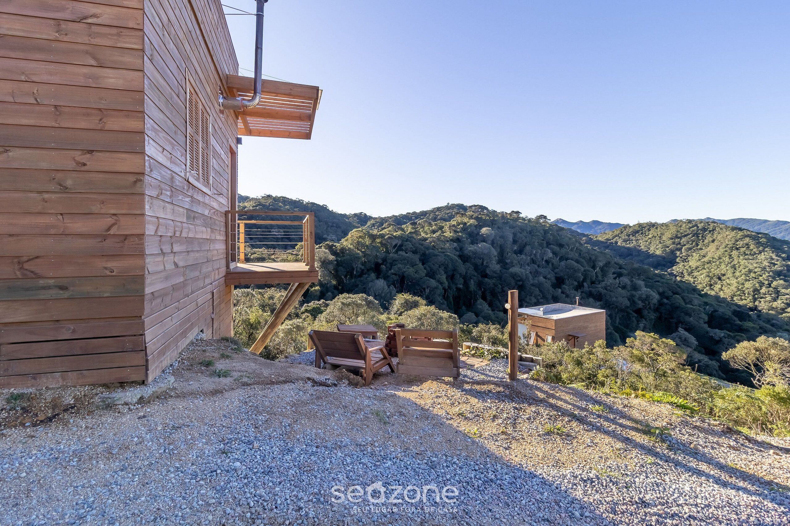 Una cabaña de madera con un patio de grava, bancos de madera y vistas a una ladera boscosa.