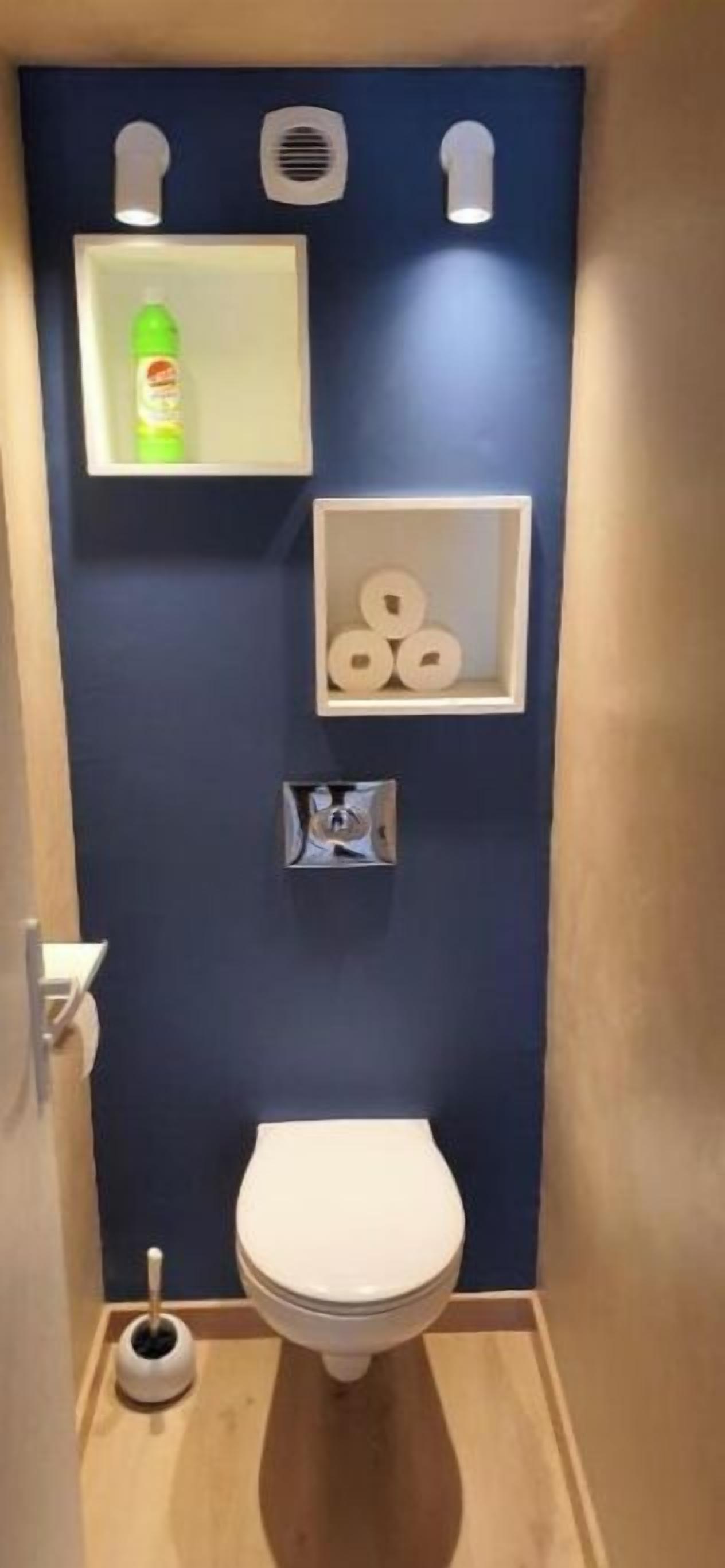 Um banheiro pequeno com uma parede azul de destaque, vaso sanitário, prateleira com papel higiênico, saboneteira e escova sanitária.