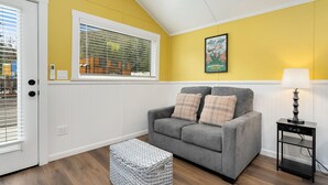 A room with a grey sofa, a small table with a lamp, a wicker ottoman, a window with blinds, and a wall-mounted poster. - Cabin 2- Youngs Gulch (Bellvue)