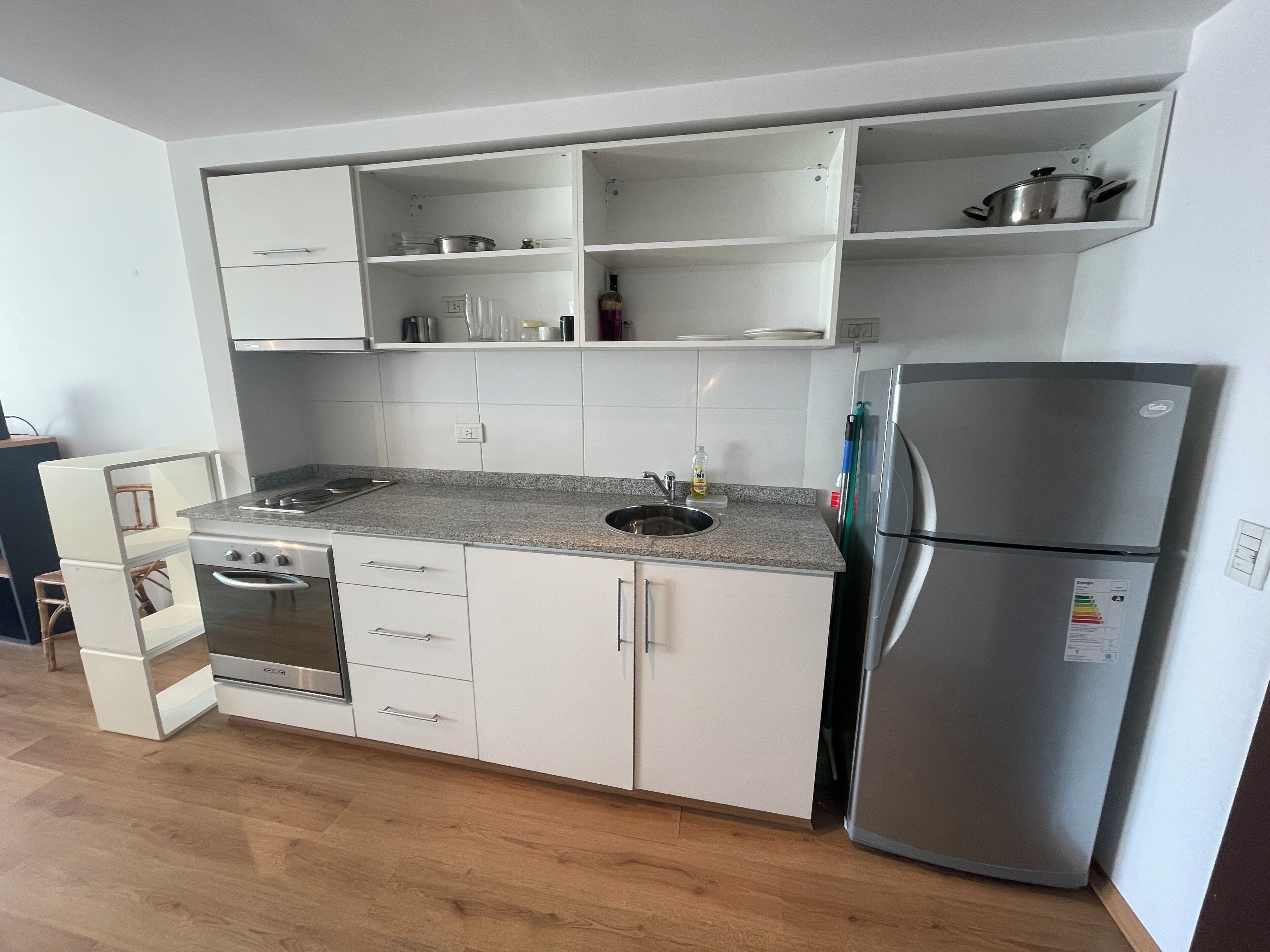 Een compacte keuken met witte kasten, een roestvrijstalen koelkast, een inbouwoven en een granieten werkblad.