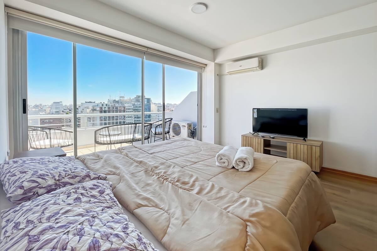 A hotel room with a large bed, a TV, and a balcony with a city view.