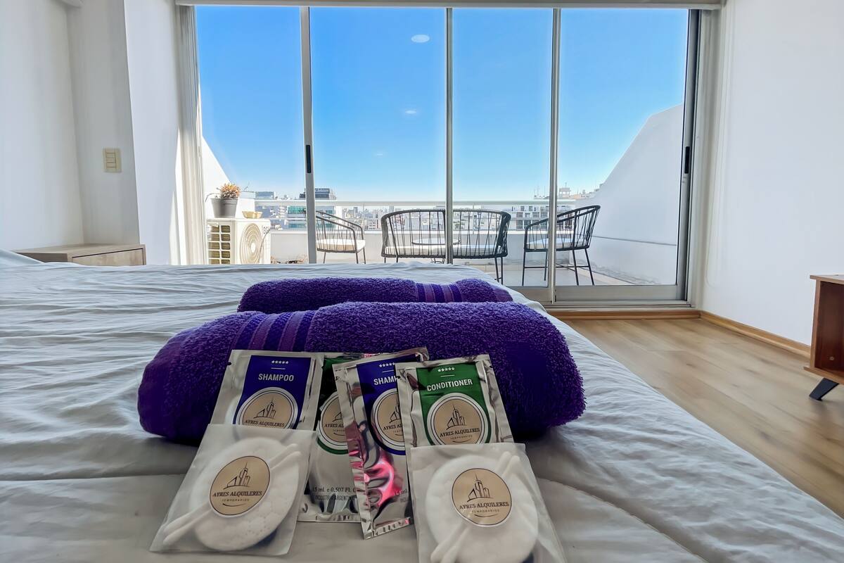 A hotel room with a large bed, a view of the city through floor-to-ceiling windows, and a balcony with chairs.