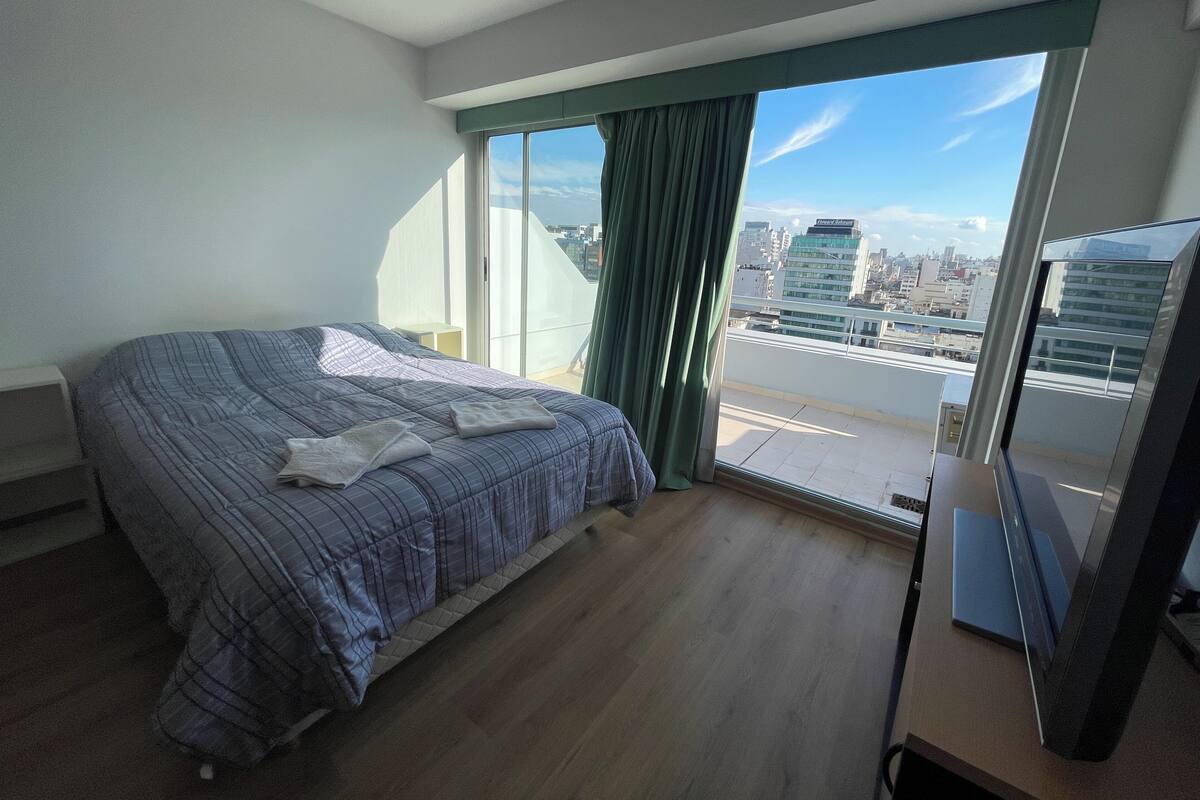 A bedroom with a large bed, a TV, and a city view through a balcony.