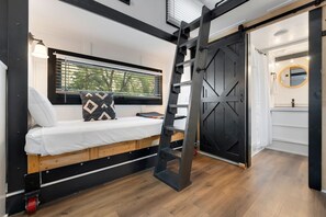 A compact bedroom with a bunk bed, a ladder, a window with blinds, and a sliding door leading to a bathroom. - Tiny 5- Profile Rock (Bellvue)