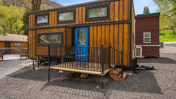 Una cabaña de madera compacta con puerta azul, una pequeña terraza y barandilla negra.
