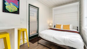 Hotellihuone, jossa on sänky, työpöytä, kaksi keltaista jakkaraa, ikkuna, jossa on sälekaihtimet, ja seinälle kiinnitetty taideteos.