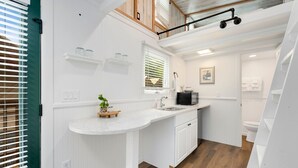 Una cucina compatta con mobili bianchi, un piano di lavoro in marmo e un piccolo isolotto. Si trova un forno a microonde, un lavandino e una finestra con persiane.