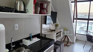 Cocina moderna con encimera negra, electrodomésticos de acero inoxidable y horno empotrado. Cuenta con un pequeño comedor con una silla y una mesa, y un amplio ventanal con vistas a la ciudad.