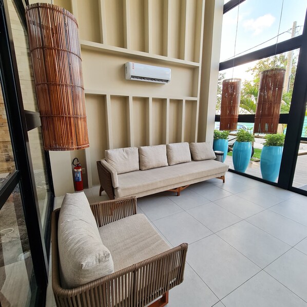 A modern lounge area with a sofa and a wicker chair, a wall-mounted air conditioner, and potted plants.