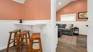 Ein Zimmer mit weißer Wandverkleidung, einer Bar mit Holzstühlen, einem grauen Sofa und einem gerahmten Bild an der Wand.