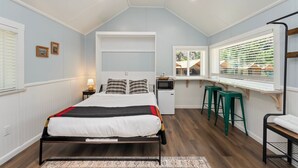 Un dormitorio con cama, escritorio y silla, ventana con persianas y una pequeña kitchenette.