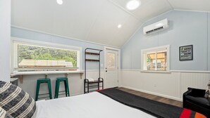 Un dormitorio con una cama, dos taburetes verdes, un tapete negro y rojo, un equipo de aire acondicionado y una ventana con persianas.