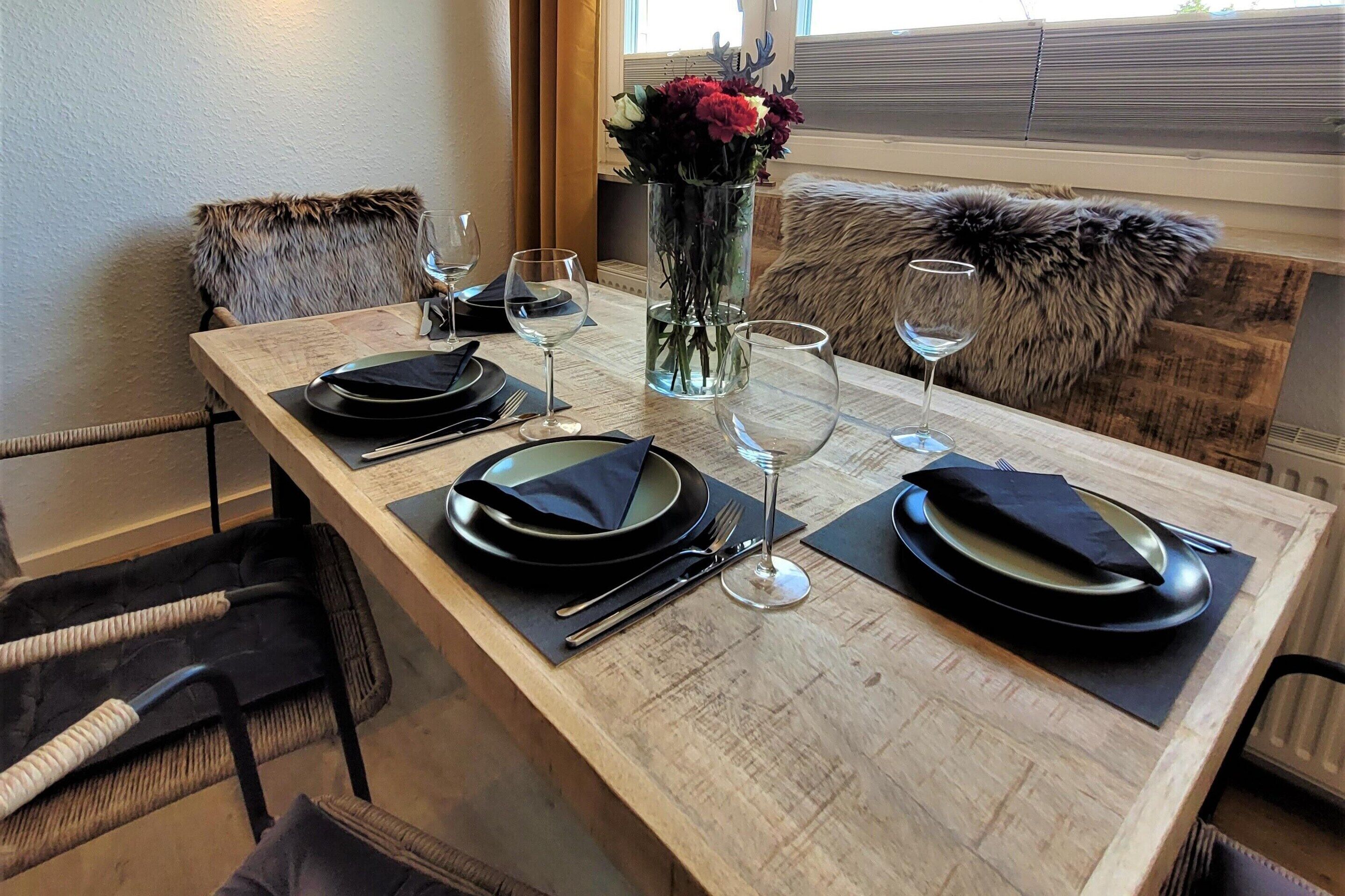 A dining table set with plates, cutlery, and wine glasses, flanked by chairs with fur throw pillows.