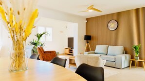 A modern living room with a wooden accent wall, a dining area with a vase of dried flowers, and a ceiling fan.