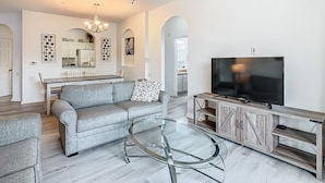 A modern living room with a grey sofa, a glass coffee table, a TV on a wooden stand, and a ceiling fan. - 3BR Disney Condo | Water Slides Fun (Kissimmee)