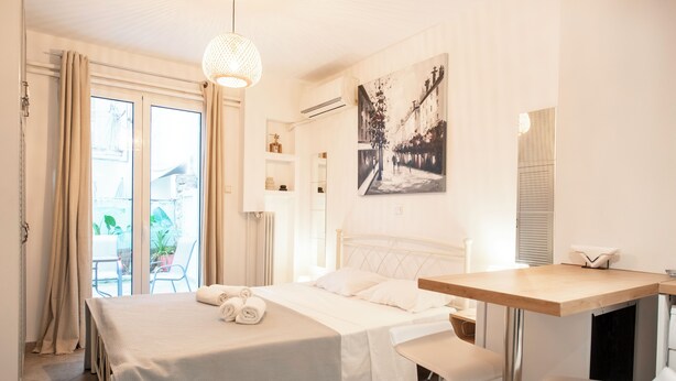 Une chambre d’hôtel avec un lit, un bureau et une chaise. Il y a une fenêtre avec des rideaux, un tableau au mur et une plante en pot à l’extérieur.