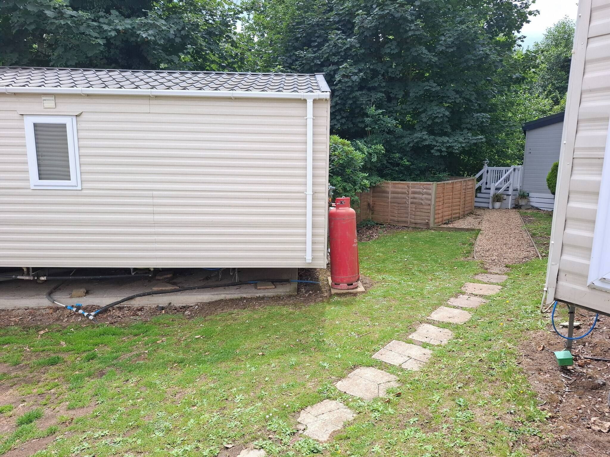 A mobile home with a small attached garage, a gas cylinder, and a paved pathway leading to a wooden fence.