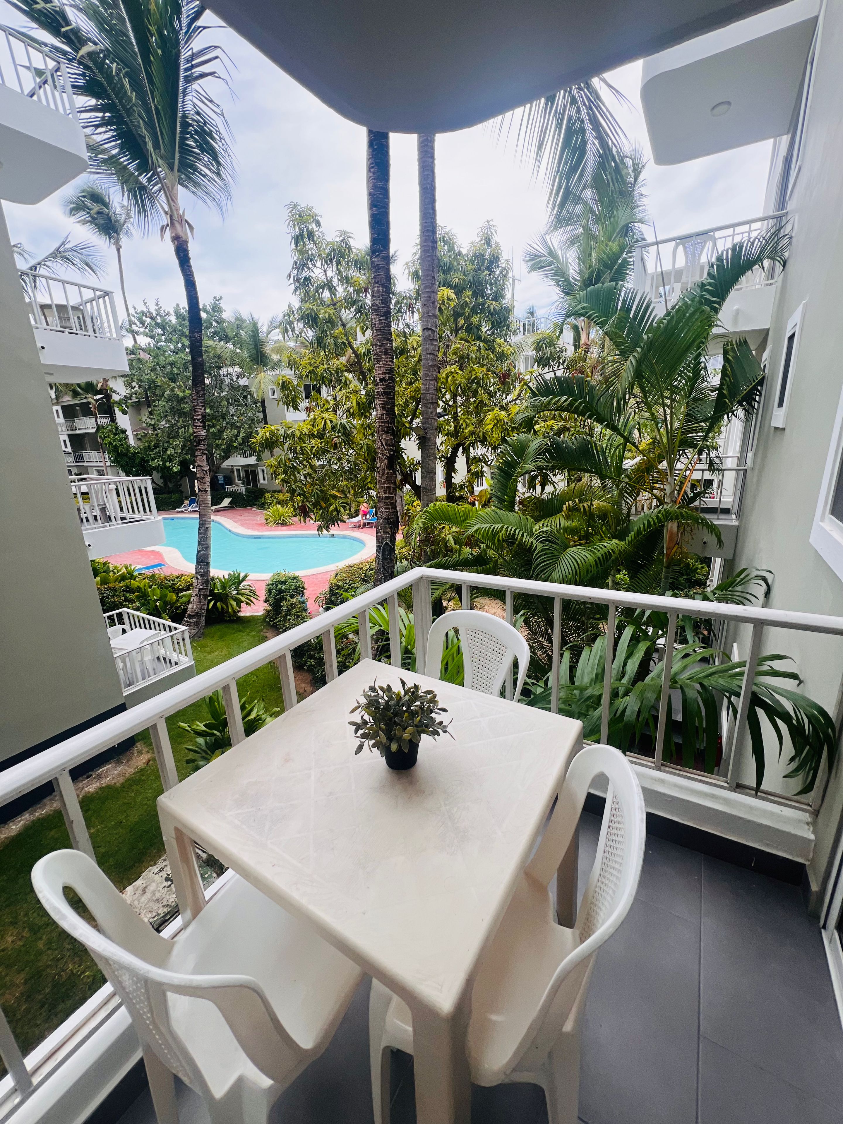 A balcony with a table and chairs, overlooking a swimming pool and lush greenery.