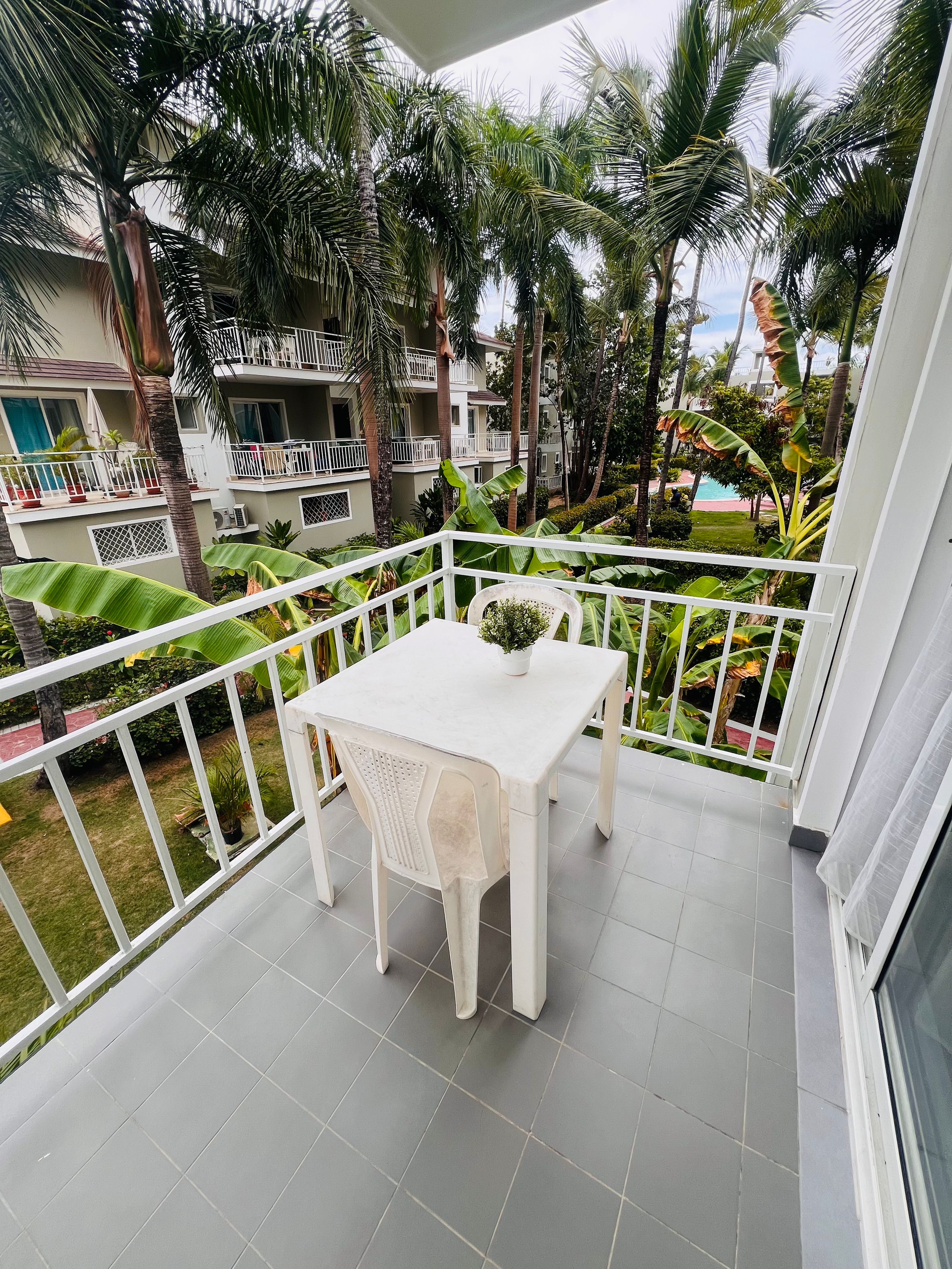 Een balkon met een witte tafel en stoel, omgeven door een witte reling, met uitzicht op een zwembad en weelderige begroeiing.
