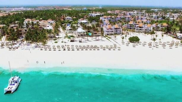 An aerial view of a coastal resort with beachfront villas, a sandy beach, and a boat docked near the shore. - Ducassi Rooftop Pool Beach Suites (Punta Cana)