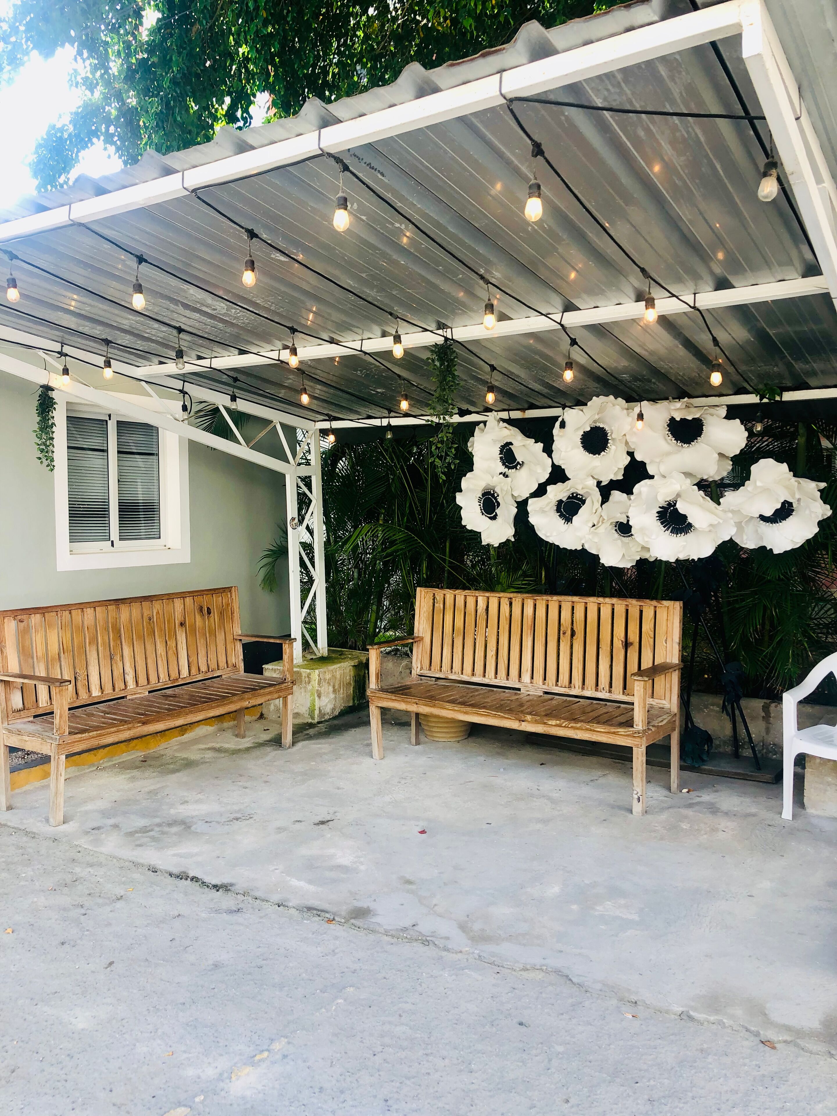 An outdoor seating area with two wooden benches under a metal roof, surrounded by greenery.
