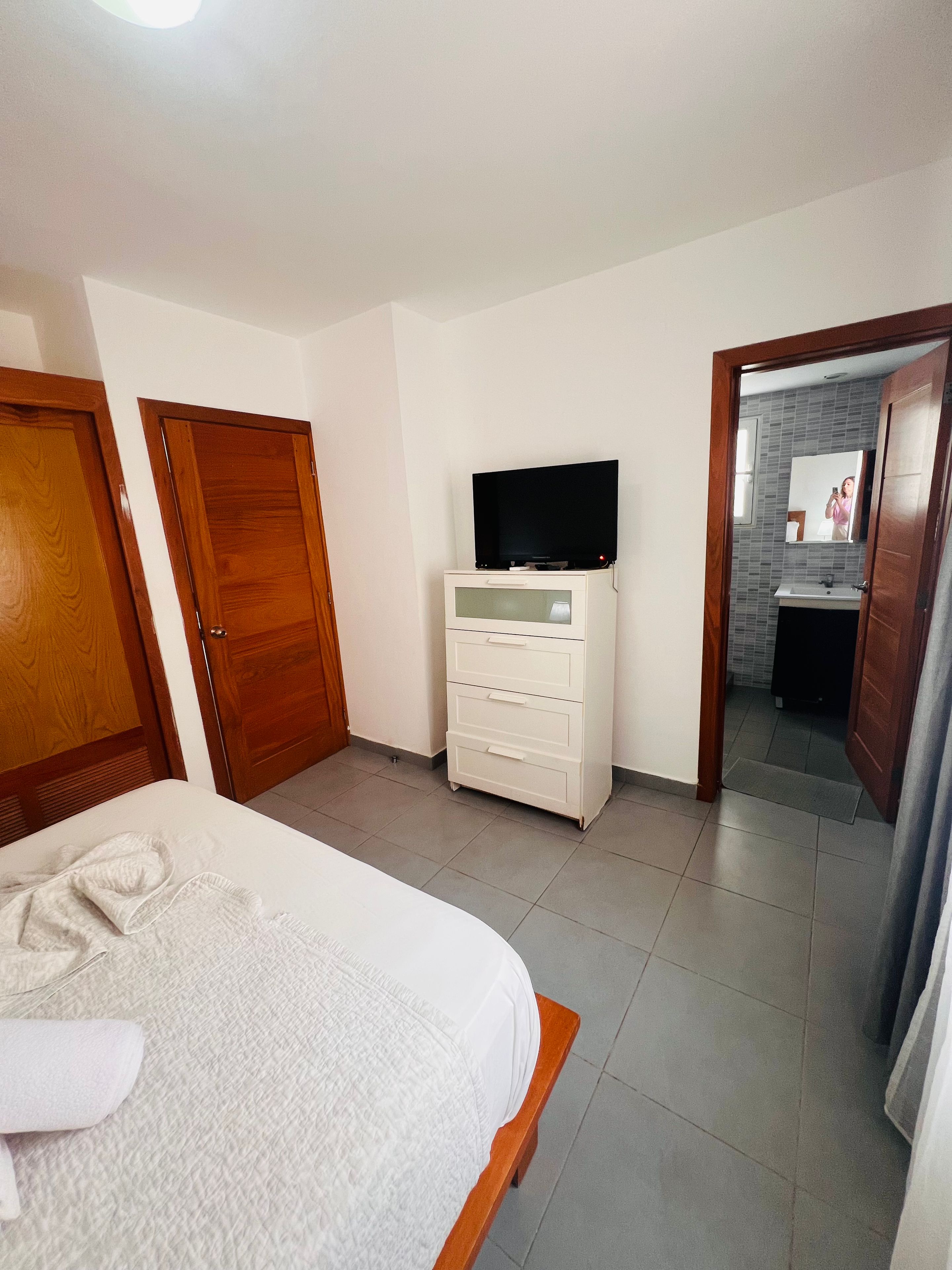 A bedroom with a bed, a dresser, a television, and a bathroom visible through an open door.