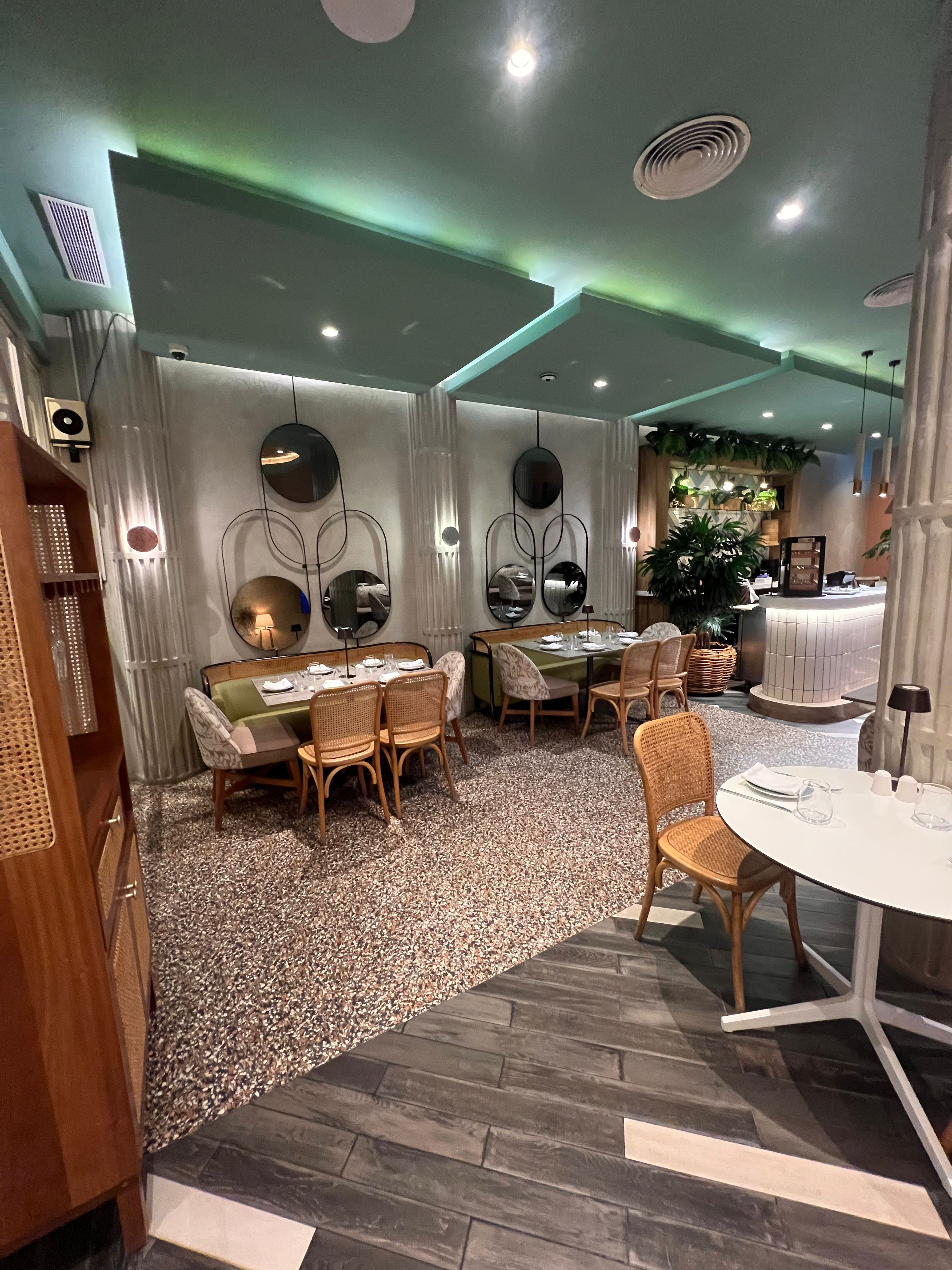 Spiseområde med runde borde og fletstole, et receptionsskrank og en plante i baggrunden.