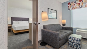 A hotel room with a bed, a sofa, a patterned ottoman, a floor lamp, a wall-mounted picture, and a radiator. - Home2 Suites By Hilton Lebanon (Lebanon)