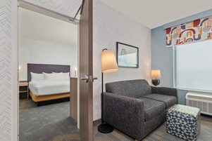A hotel room with a bed, a sofa, a patterned ottoman, a floor lamp, a wall-mounted picture, and a radiator.