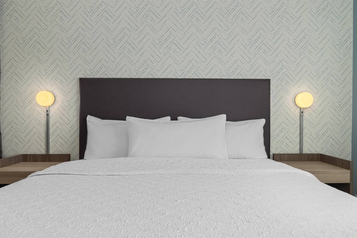 A neatly made bed with a dark headboard, two bedside lamps, and a herringbone-patterned wallpaper.
