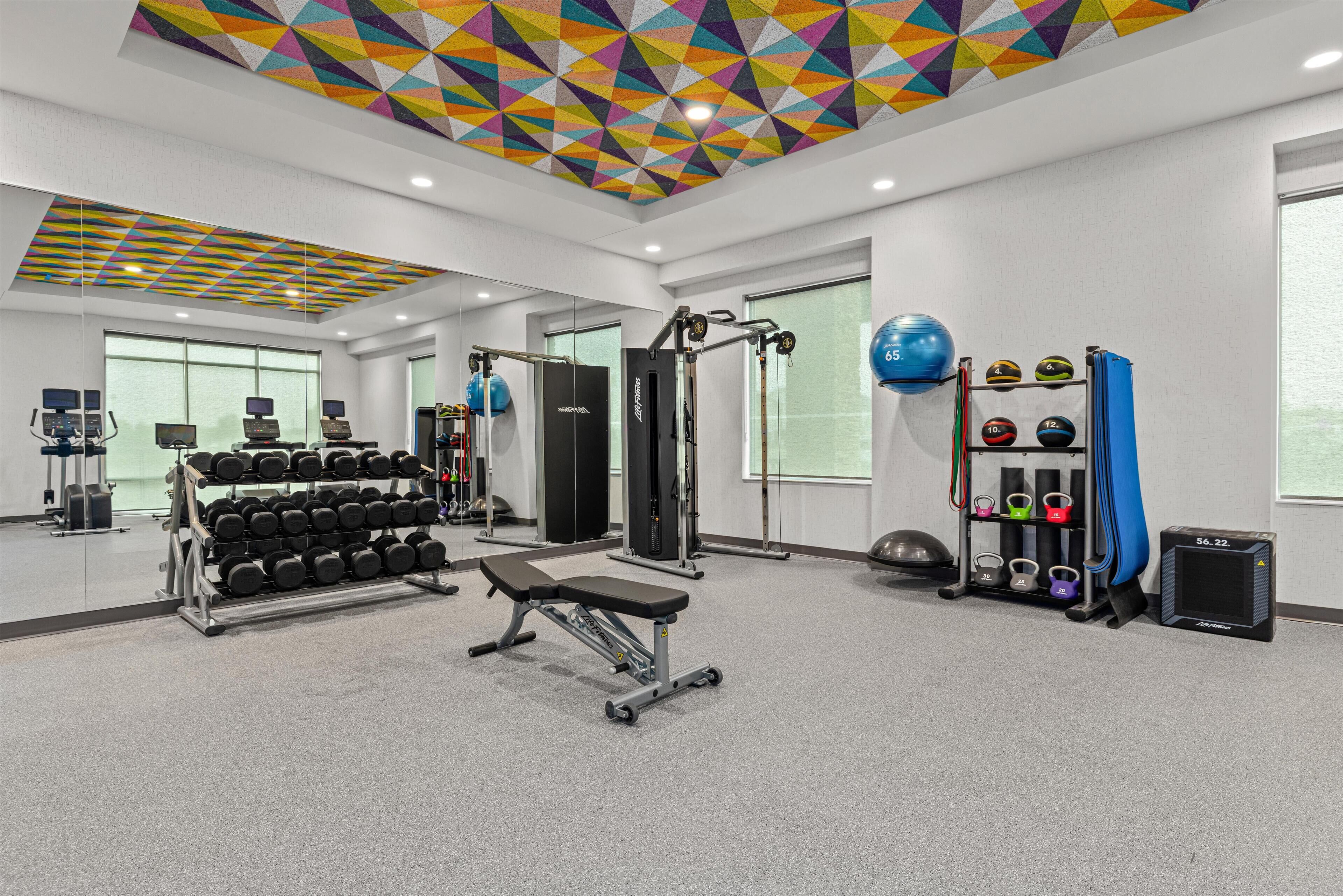 Een sportschool met trainingsapparatuur, een bank, gewichtsschijven en een plafond met geometrische patronen.