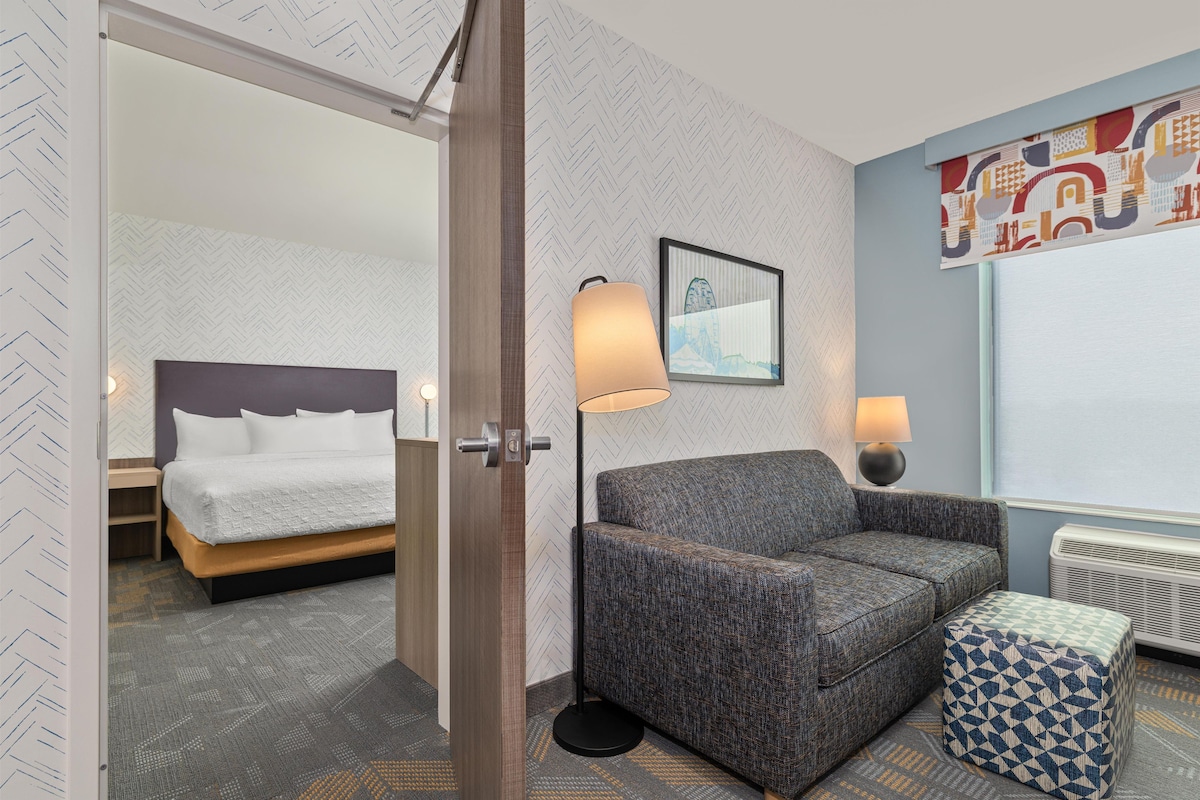 A hotel room with a bed, a sofa, a patterned ottoman, a floor lamp, a wall-mounted picture, and a radiator.