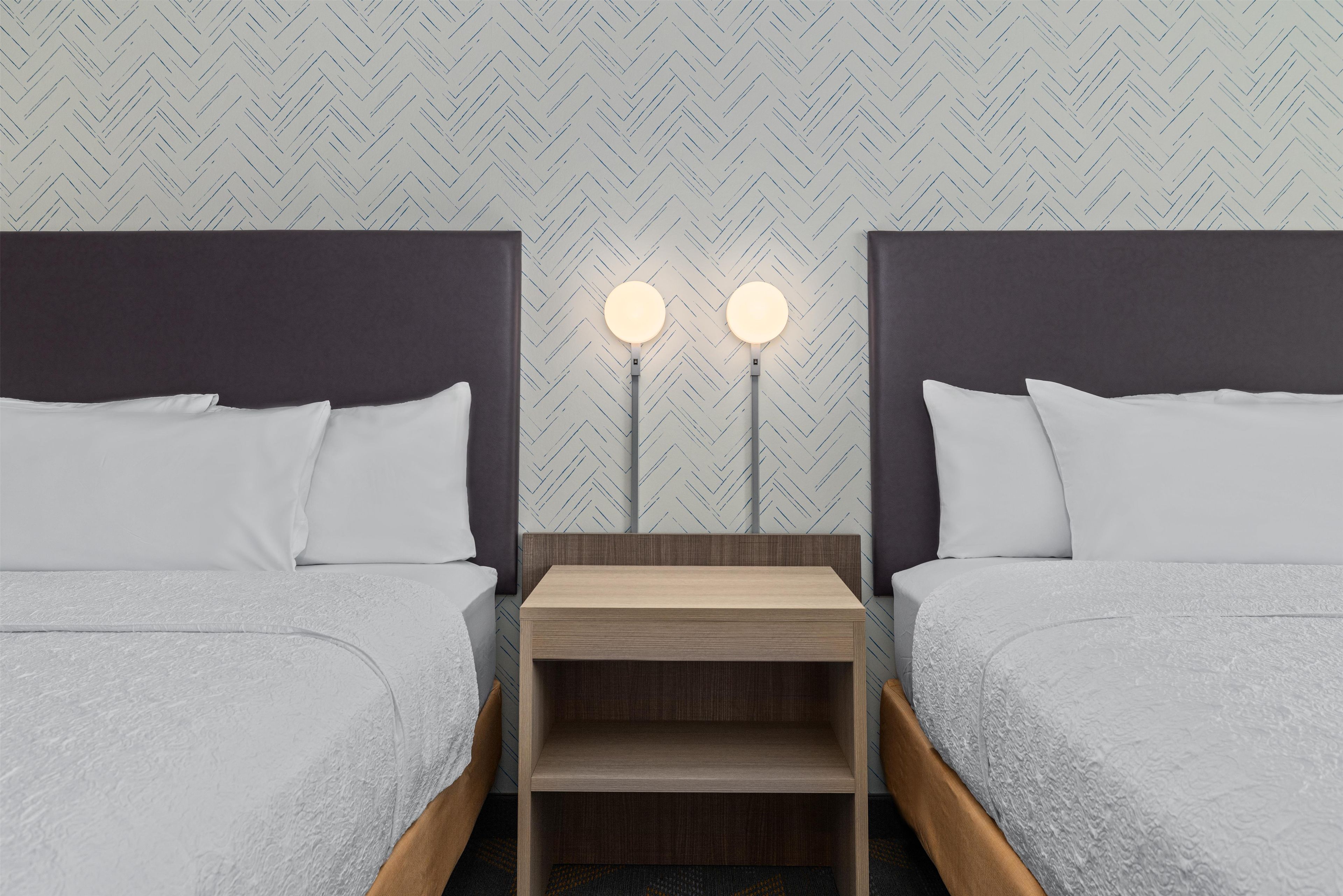 Habitación de hotel con dos camas, una mesita de noche con dos lámparas fijadas a la pared y un papel tapiz con diseño de espiga.